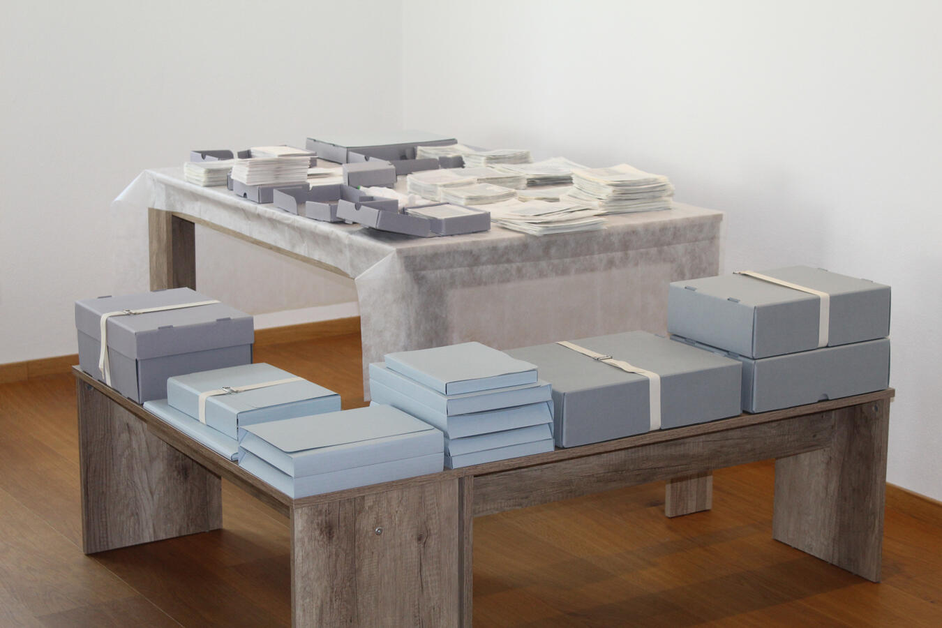 In einem hellen Raum befinden sich zwei Holztische, auf denen zahlreiche Archivmaterialien ordentlich gestapelt sind. Der Tisch ist mit einem weißen Vlies bedeckt und zeigt eine Vielzahl von Fotografien und flachen Boxen, die systematisch angeordnet sind.