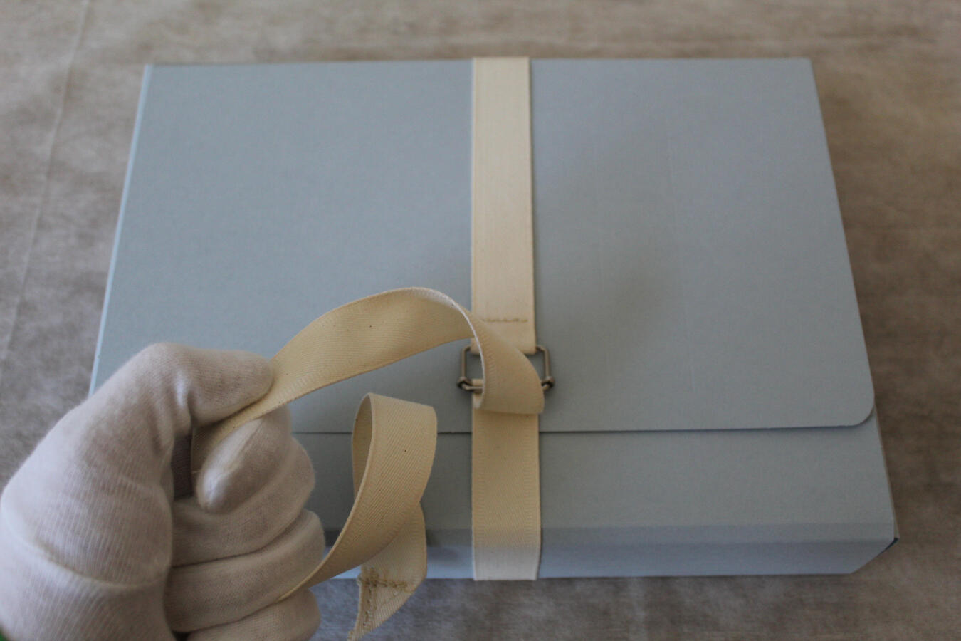 Auf dem Bild sieht man eine hellblaue Archivbox, die mit einem cremefarbenen Stoffband verschlossen ist. Eine Hand, die einen weißen Handschuh trägt, hält das Band, um die Box sicher zu transportieren.